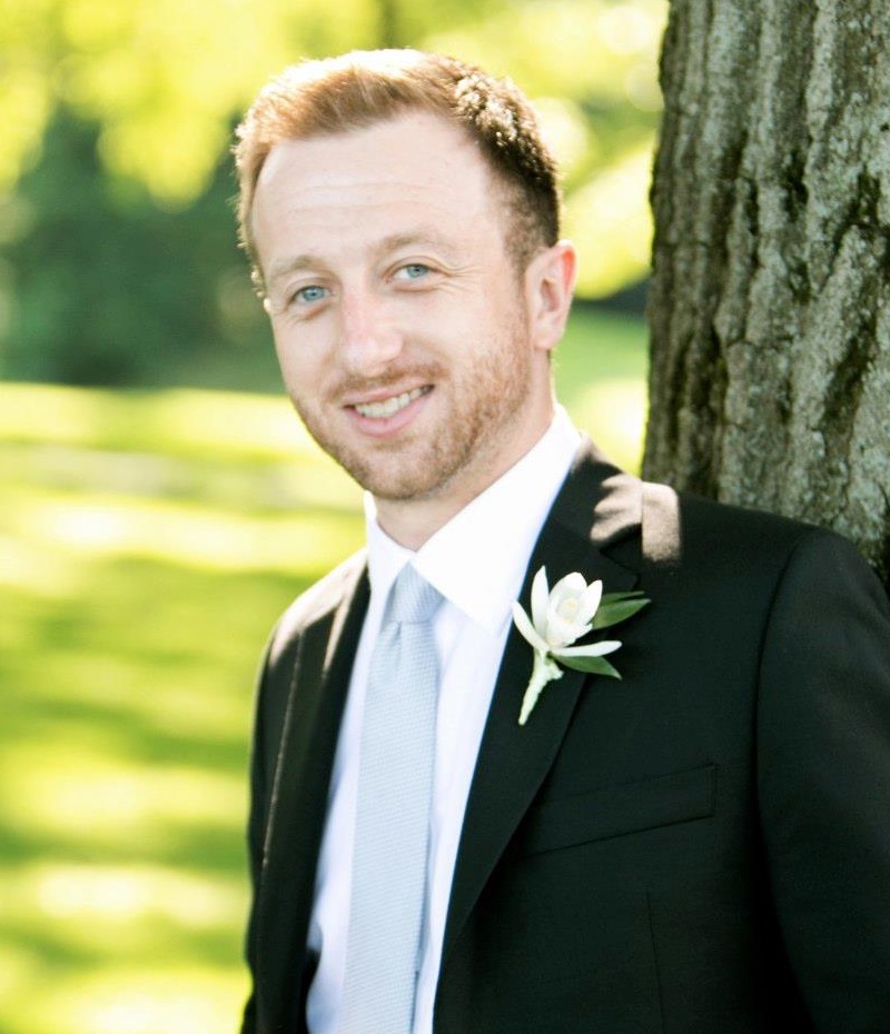 Aaron wearing a suit with a white flower on lapel, also wearing a white shirt and light blue tie.He is leaning against a tree on sunny day, smiling and looking into camera